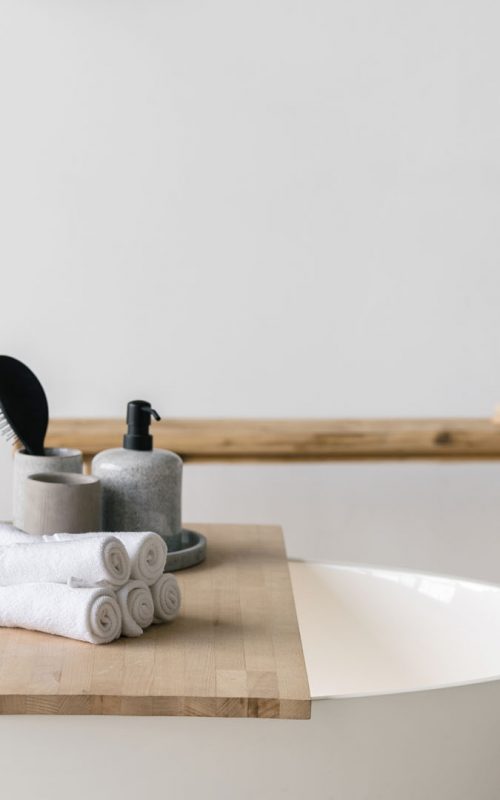 Hair brush, soap dispenser and clean fresh towels on wooden shelf over white contemporary bath with black faucet. Modern bathroom interior with bathtub and water tap